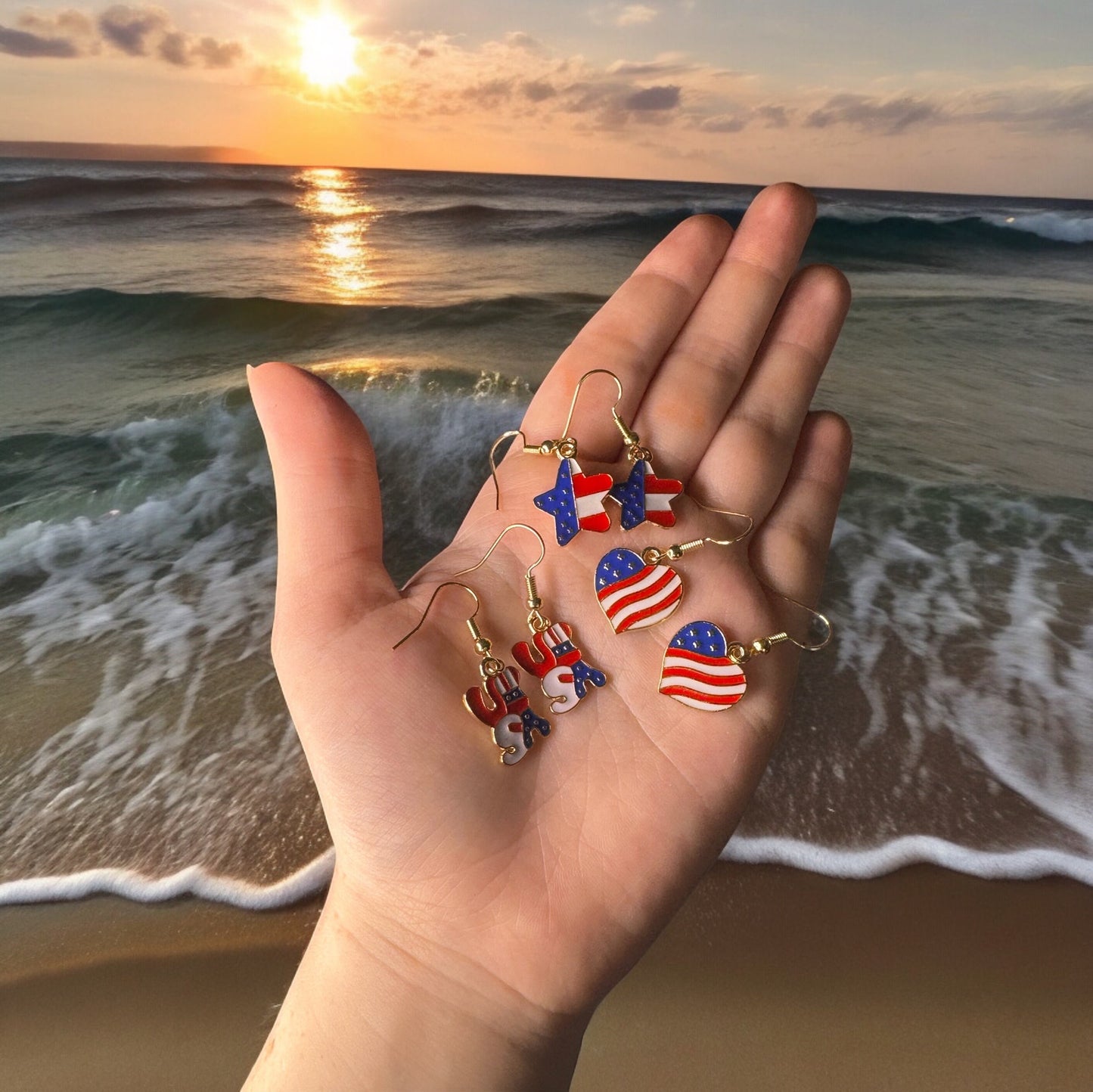 A hand holding 3 sets of 4th of July themed dangle earrings. All are printed like the U.S Flag. One is heart shaped, one is shaped in bubble letters spelling USA, and one is star shaped.