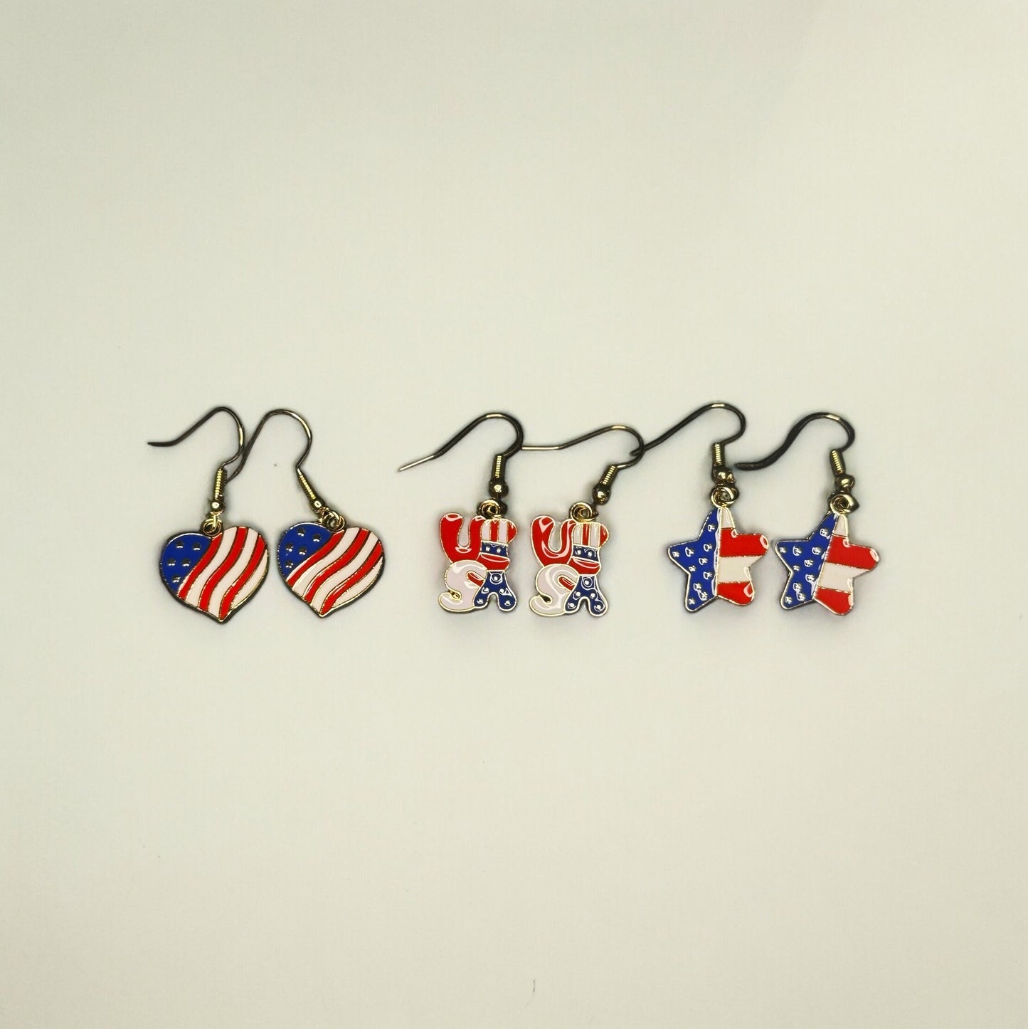 3 sets of 4th of July themed dangle earrings. All are printed like the U.S Flag. One is heart shaped, one is shaped in bubble letters spelling USA, and one is star shaped.