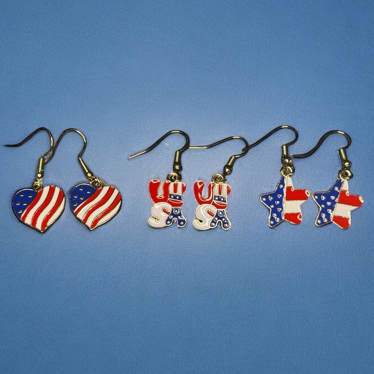 3 sets of 4th of July themed dangle earrings. All are printed like the U.S Flag. One is heart shaped, one is shaped in bubble letters spelling USA, and one is star shaped.