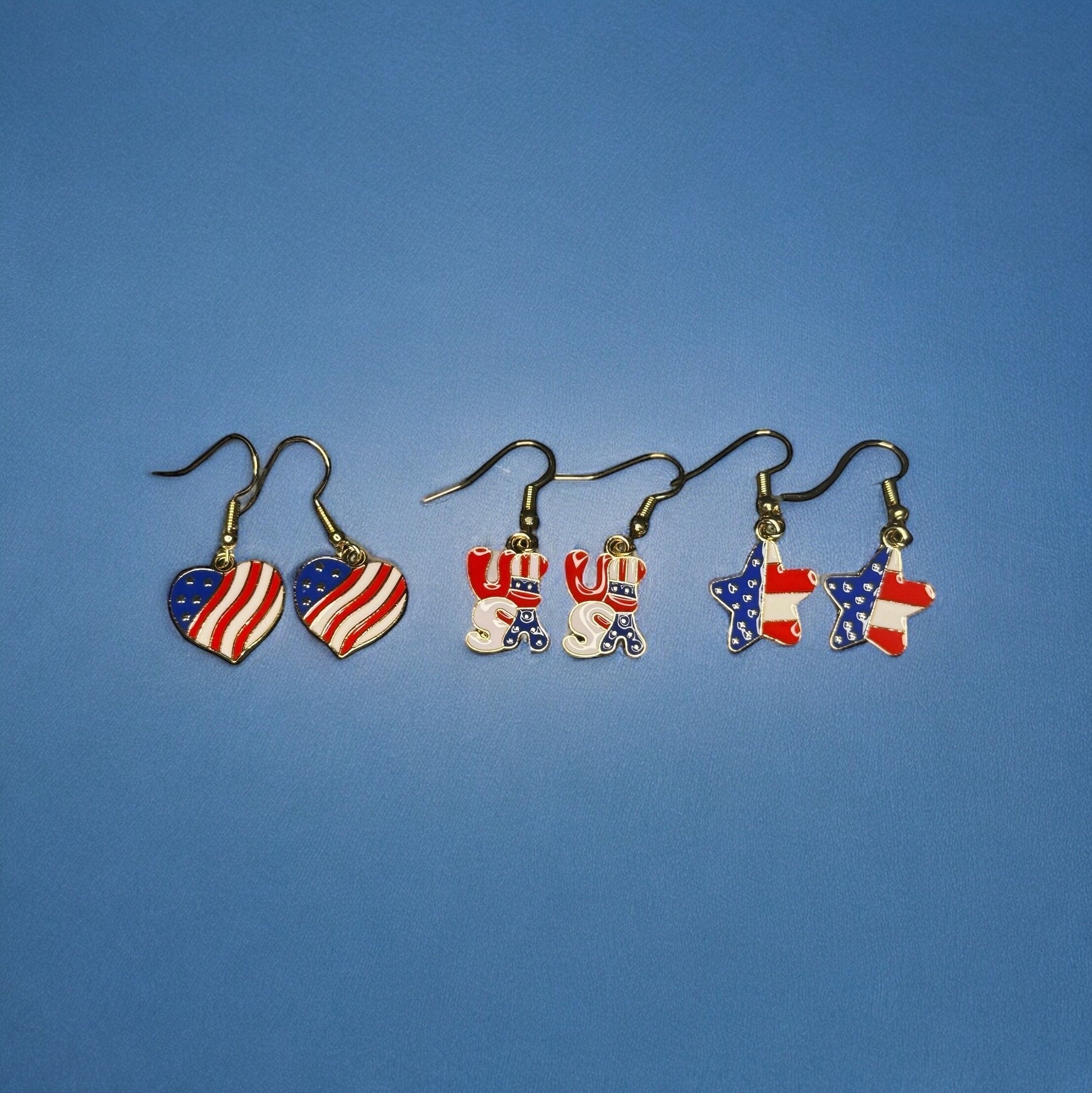 3 sets of 4th of July themed dangle earrings. All are printed like the U.S Flag. One is heart shaped, one is shaped in bubble letters spelling USA, and one is star shaped.