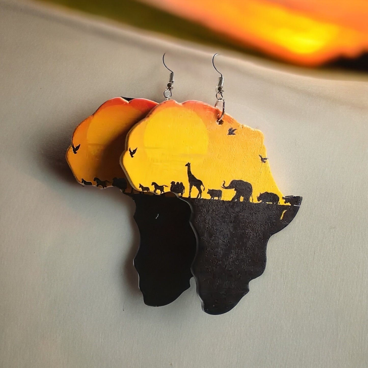 A pair of dangle earrings shaped like the continent of Africa depicting a warm orange and yellow sunset and a black silhouette of various African animals walking in a line on a pasture silhouette.