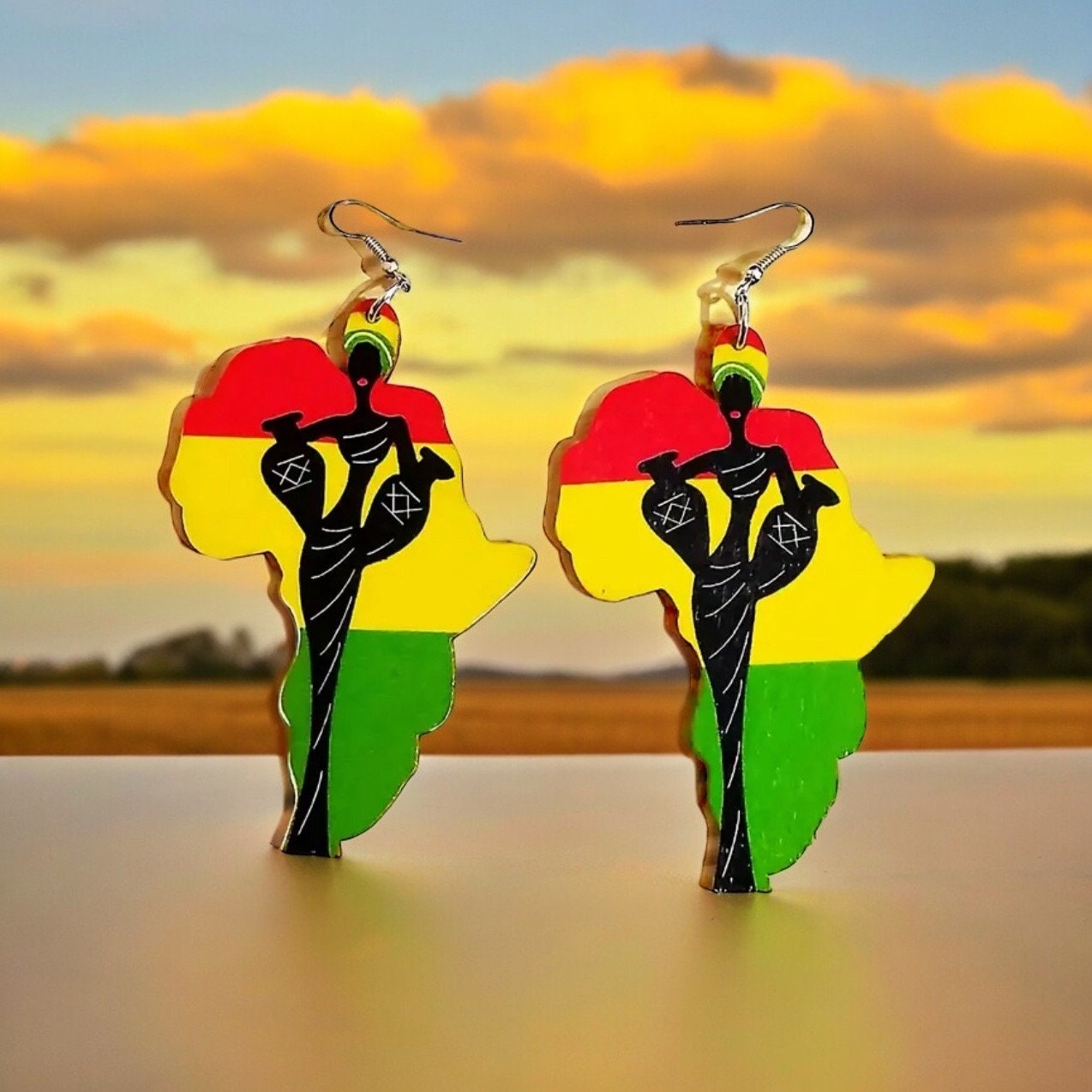 A pair of dangle earrings shaped like the continent of Africa with a red, yellow and green Ghana flag print in the background and a black silhouette of a beautiful African woman holding two vases and wearing a head covering on the front.