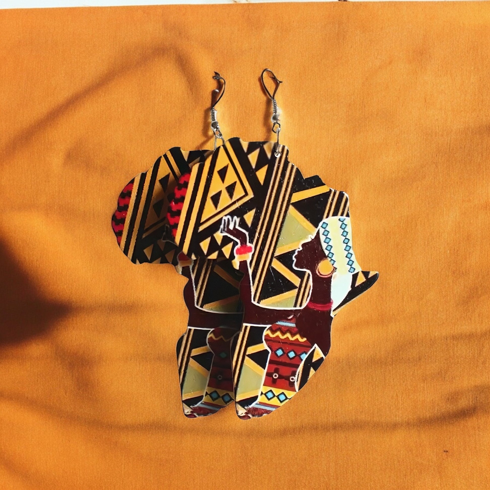 A pair of dangle earrings shaped like the continent of Africa with a yellow and black tribal print in the background and a beautiful African woman in front of it wearing a tribal print dress, colorful bangles and earrings and a white turban.