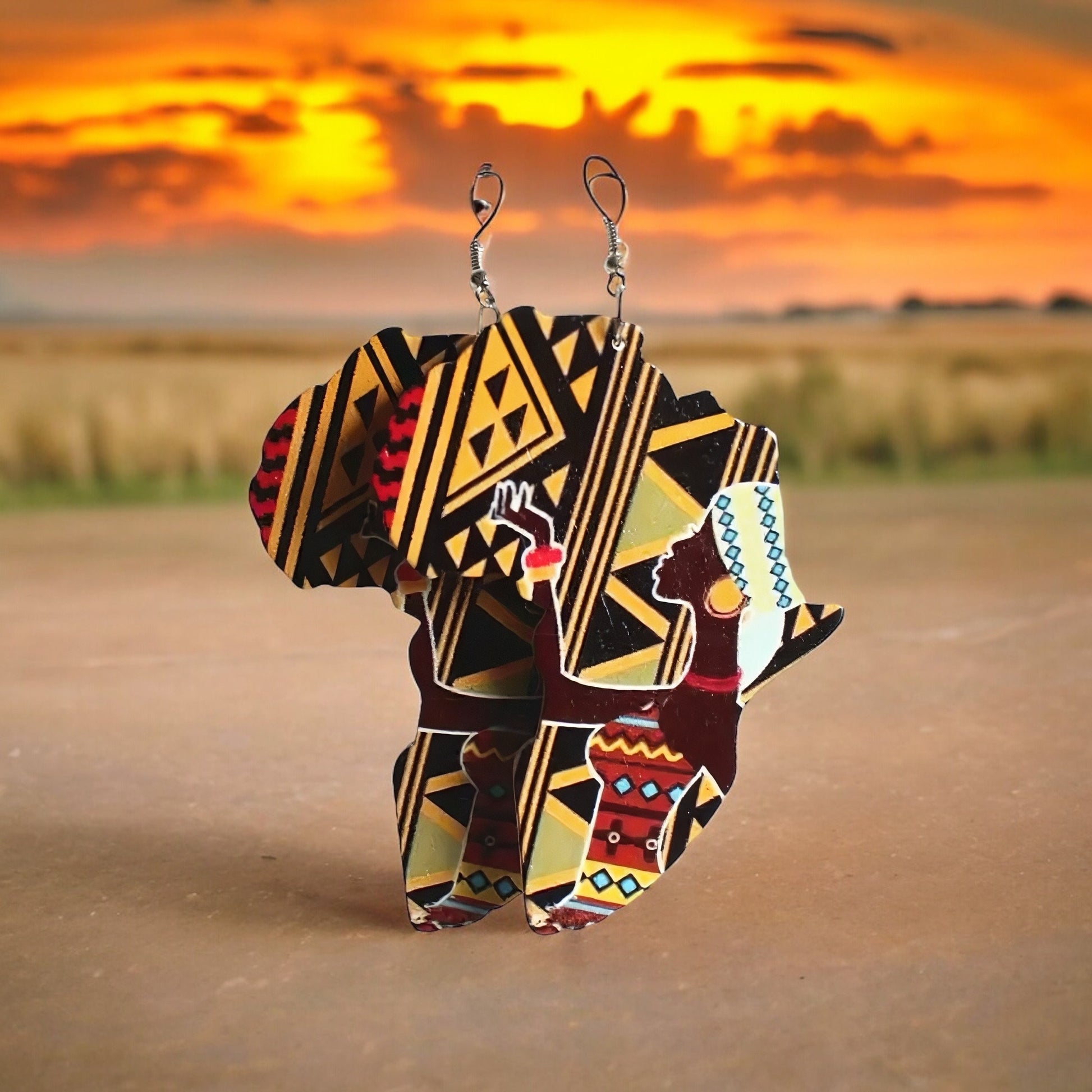 A pair of dangle earrings shaped like the continent of Africa with a black and yellow tribal print background and a beautiful African woman on the front wearing a tribal print dress, some jewelry and a head covering.