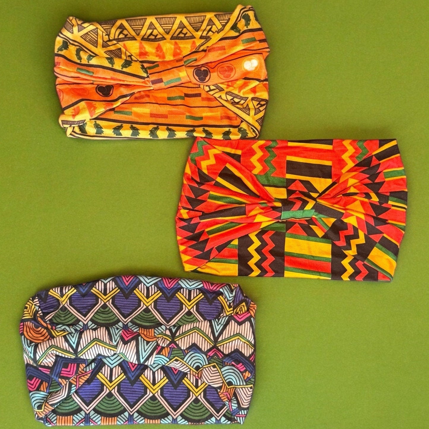 Three stretchy headbands laying on a green surface. One is an orange African tribal print, one is a red, yellow, green and black African flag style print, and the other is a blue artistic African tribal print.