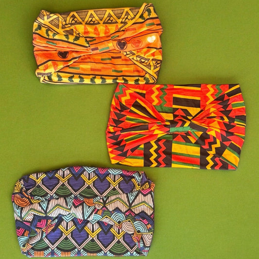 Three stretchy headbands laying on a green surface. One is an orange African tribal print, one is a red, yellow, green and black African flag style print, and the other is a blue artistic African tribal print.