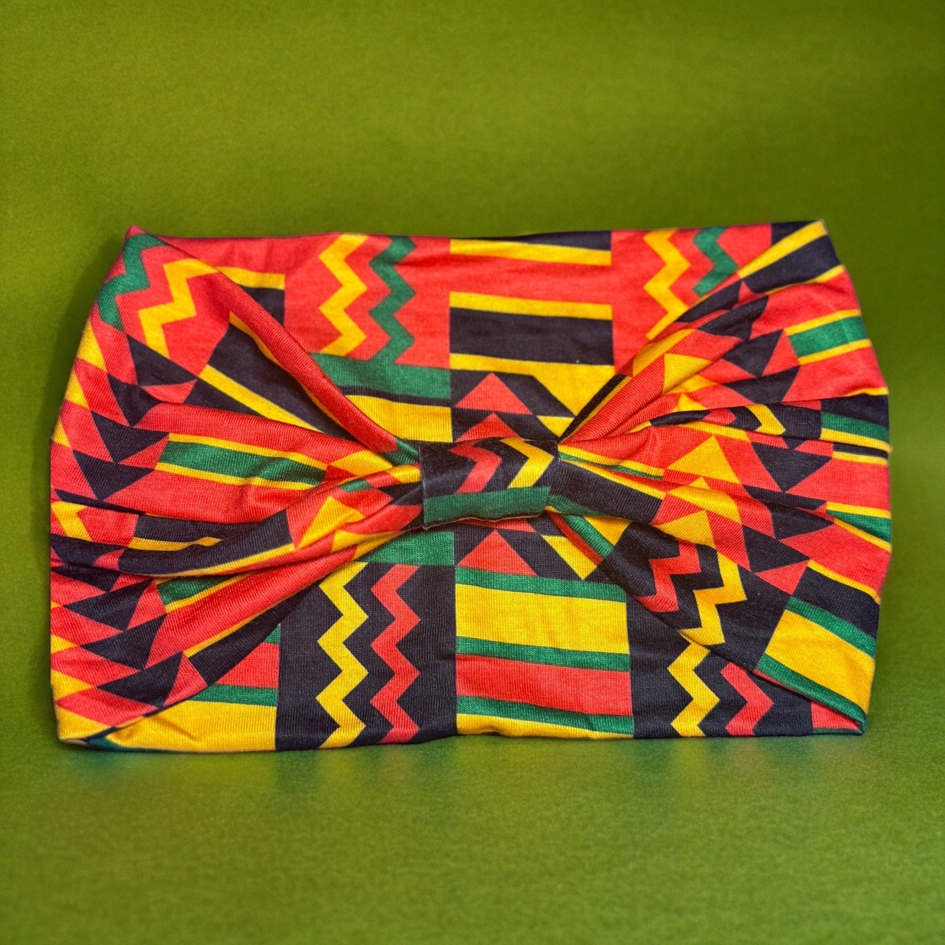 The backside of a stretchy polyester African themed headband that is colored red, yellow, green and black with an artistic tribal style print on it. The photo displays a knotted style on the headband.