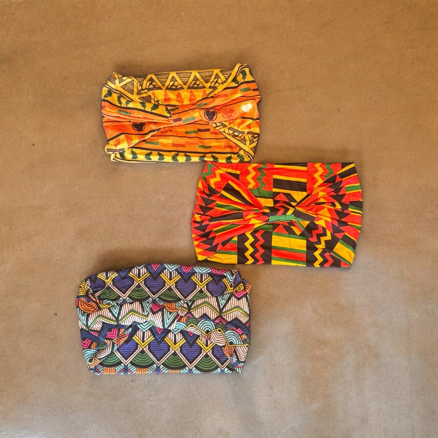 Three stretchy polyester African tribal print headbands in various colors laying on a neutral colored brown-tone background.