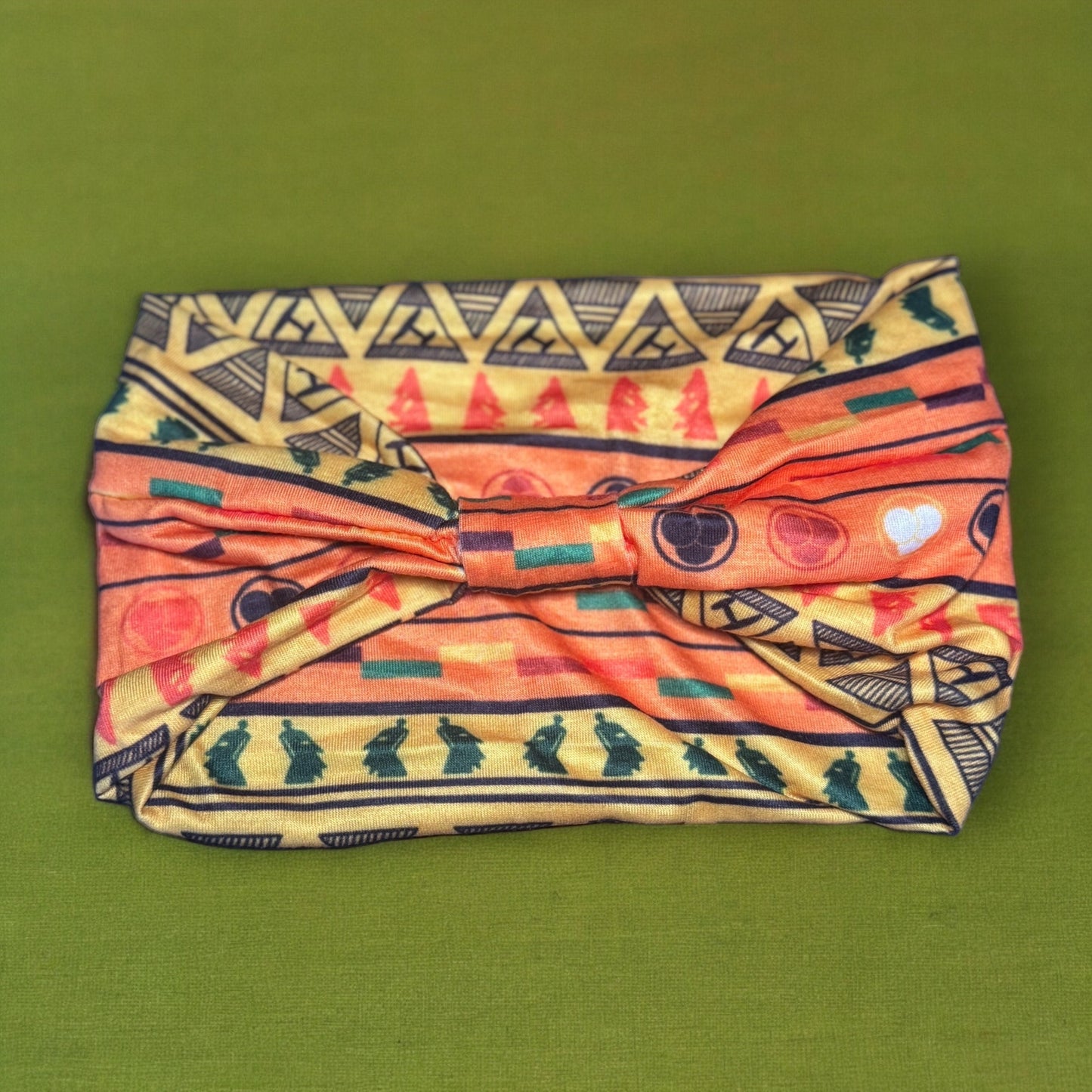 The backside of a stretchy African tribal print headband showing a knot design. The print of the headband is orange, yellow, green, black, with small amounts of white and red.