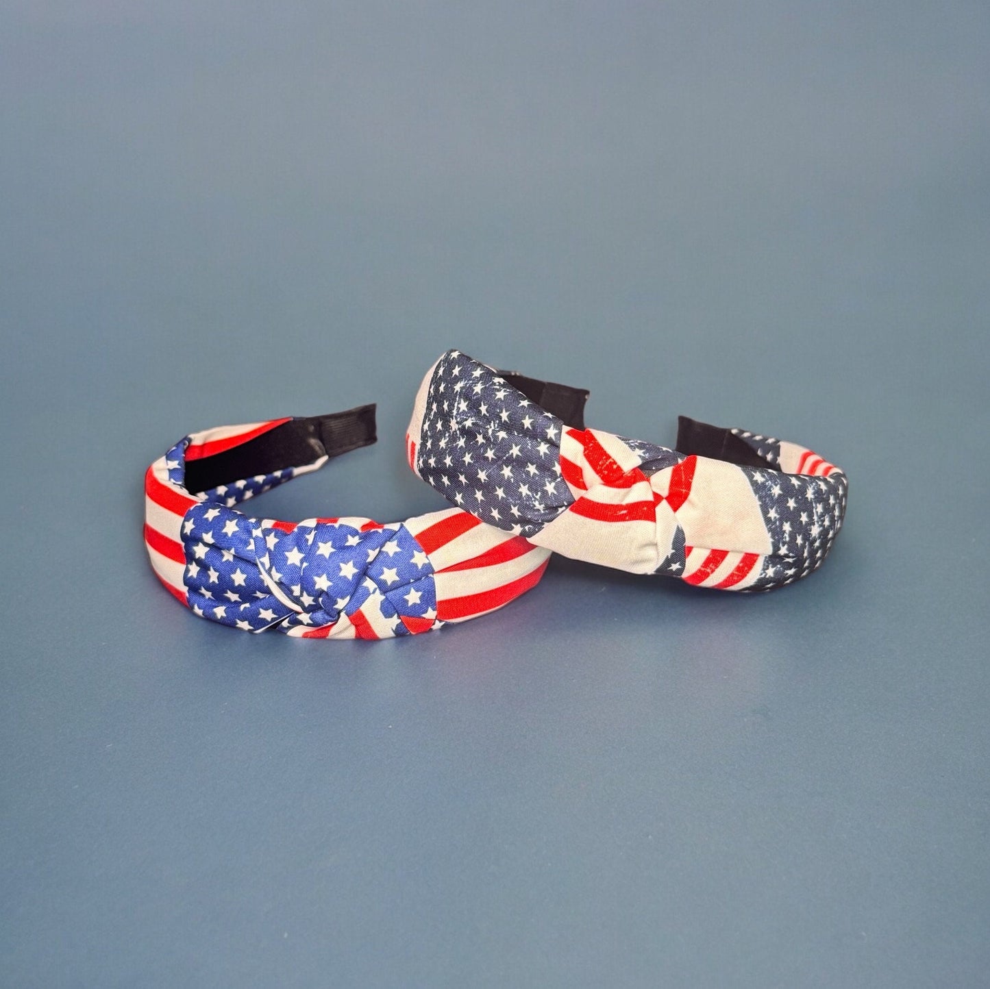 Two American flag print wide fabric headbands laying on top of each other laying on a blue surface.