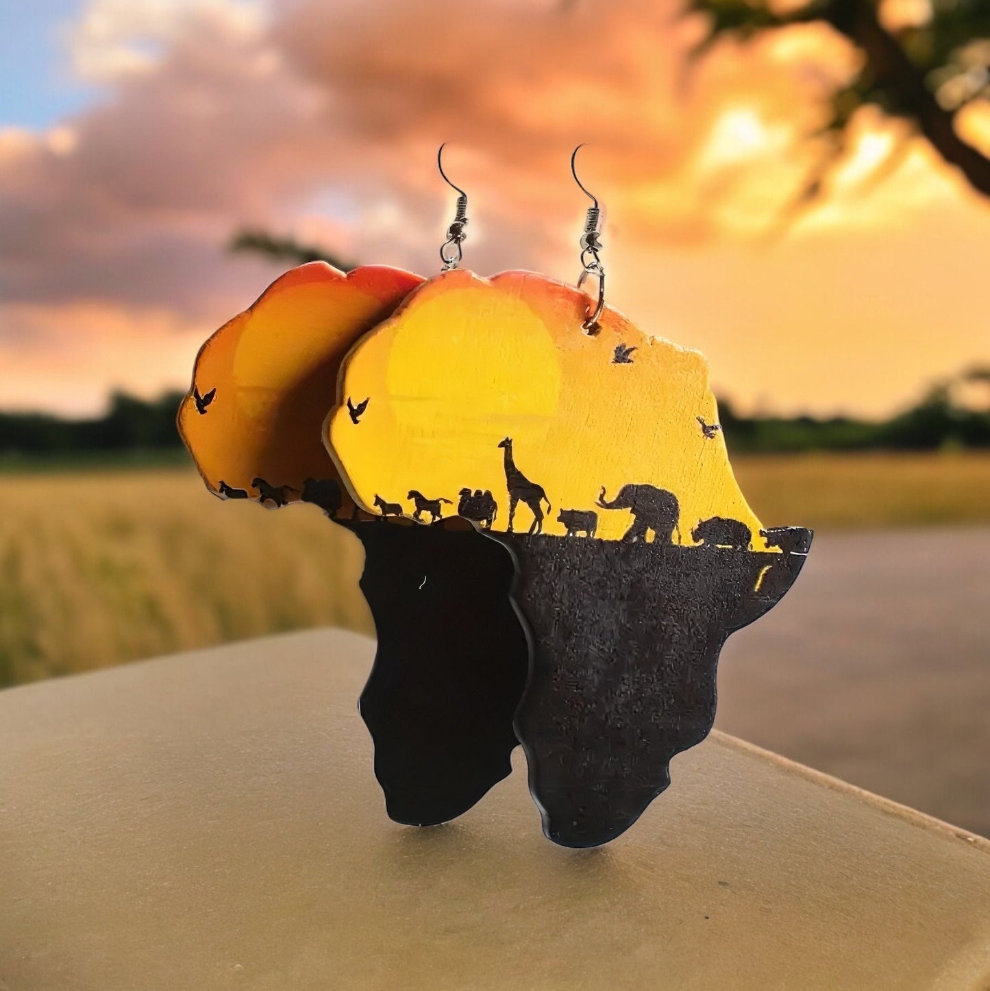 A pair of dangle earrings shaped like the continent of Africa with a warm orange and yellow sunset and a black silhouette of a pasture with various African animals such as elephants and giraffes.