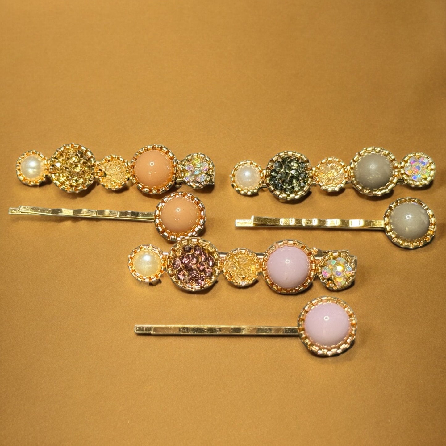 Three sets of barrettes laying on a gold colored background. Each barrette is gold with varying colors of rhinestones, colored pearls and faux pearls. One pack is peach colored, one is grey, and one is a light baby pink. One in each pair is shaped like 5 circles--2 big, 3 small--and the other is shaped like a bobby pin with one colored pearl at the end.