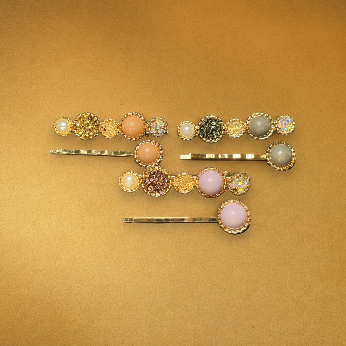 Three sets of barrettes laying on a gold colored background. Each barrette is gold with varying colors of rhinestones, colored pearls and faux pearls. One pack is peach colored, one is grey, and one is a light baby pink. One in each pair is shaped like 5 circles--2 big, 3 small--and the other is shaped like a bobby pin with one colored pearl at the end.