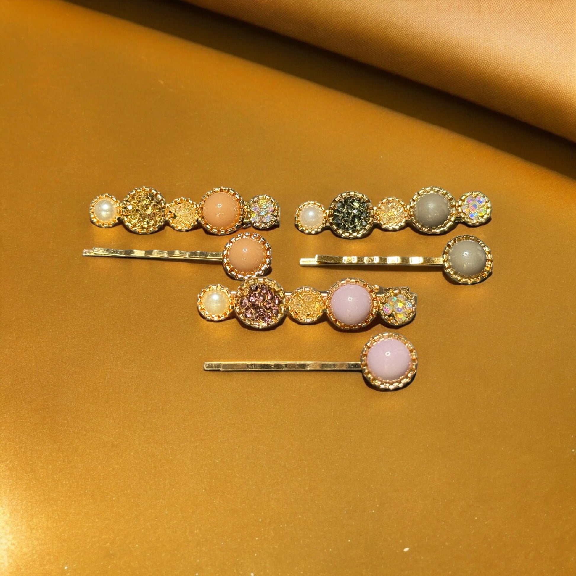 Three sets of barrettes laying on a gold colored background. Each barrette is gold with varying colors of rhinestones, colored pearls and faux pearls. One pack is peach colored, one is grey, and one is a light baby pink. One in each pair is shaped like 5 circles--2 big, 3 small--and the other is shaped like a bobby pin with one colored pearl at the end.