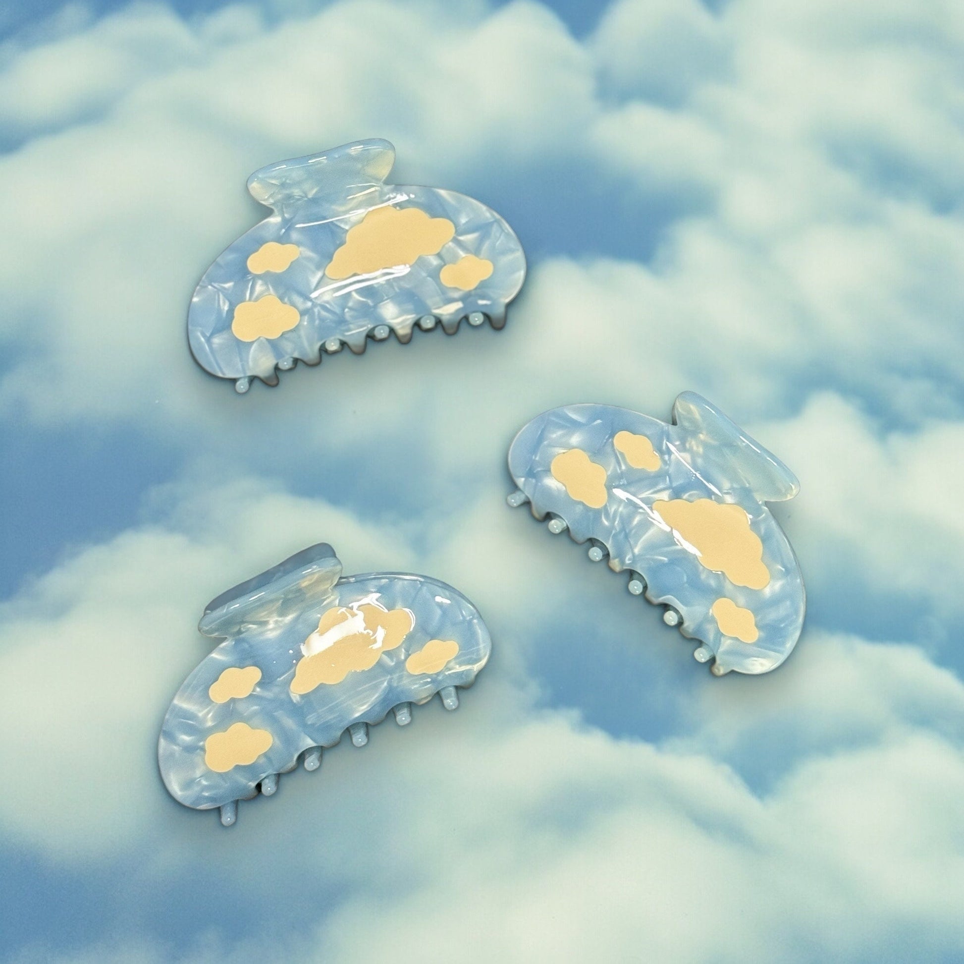 Three light blue claw clips with white clouds on them laying down on a fake cloud background.