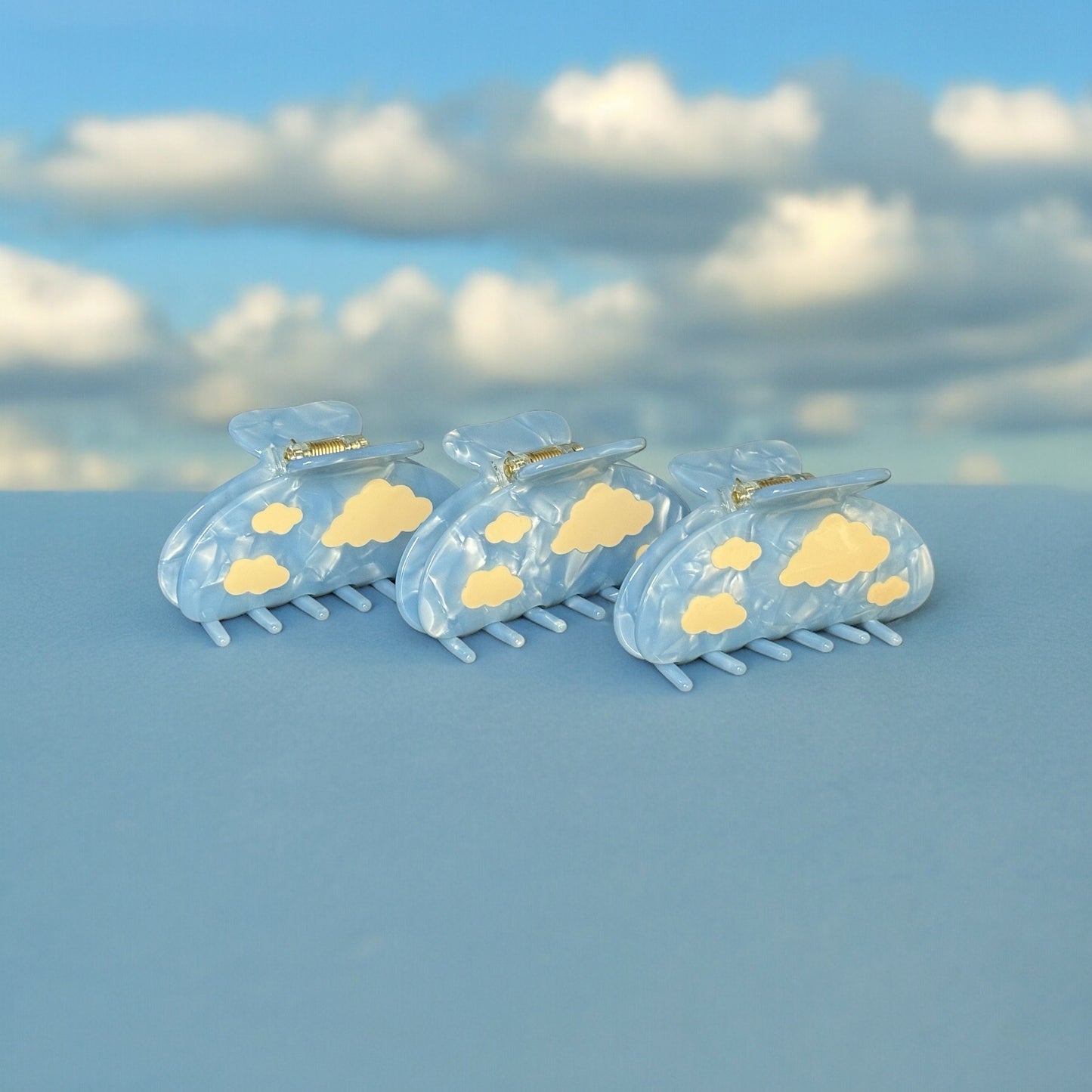 Three light blue claw clips with white clouds standing upright and at an angle on a fake cloud background.