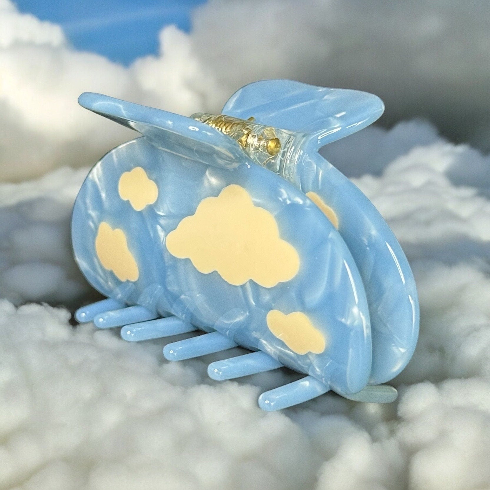 0ne light blue colored claw clip with white clouds on it standing upright but angled to the left on a fake cloud background. There is gold hardware exposed on the top that is the mechanism that opens and closes the clip.