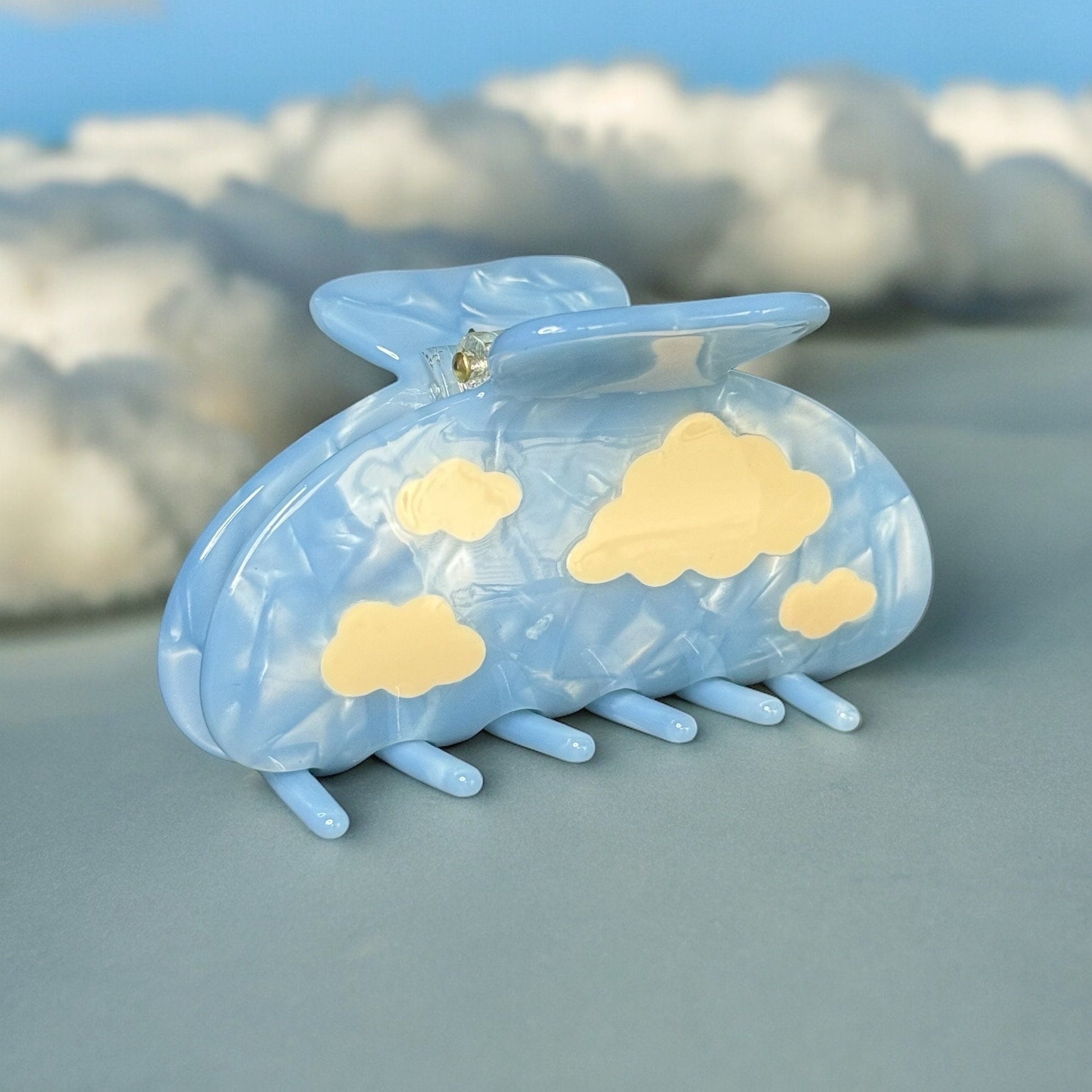 One light blue colored claw clip with white clouds on it standing upright on a fake cloud background.