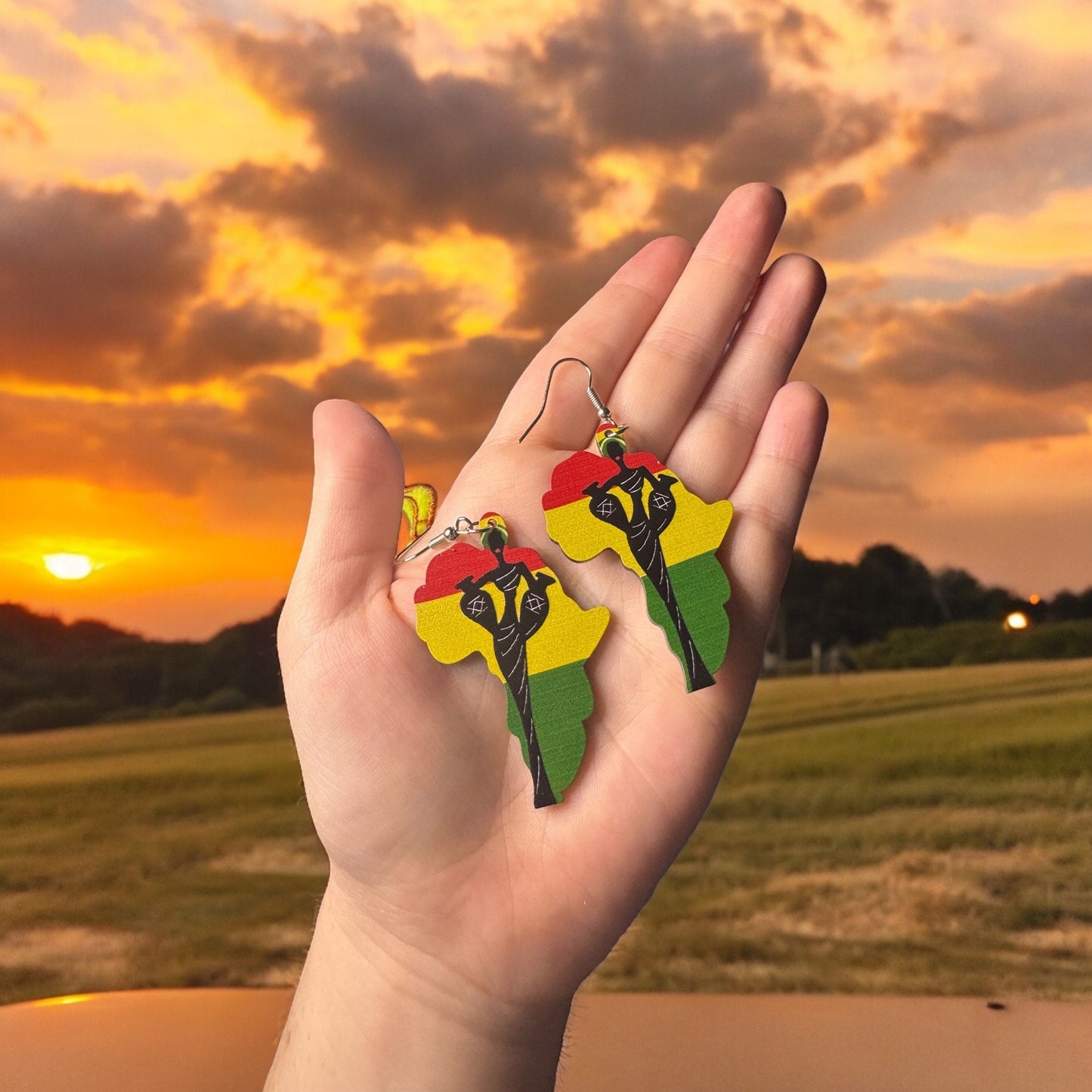 A hand holding a pair of dangle earrings shaped like the continent of Africa with a red, yellow and green flag print and the silhouette of a beautiful African woman on the front.