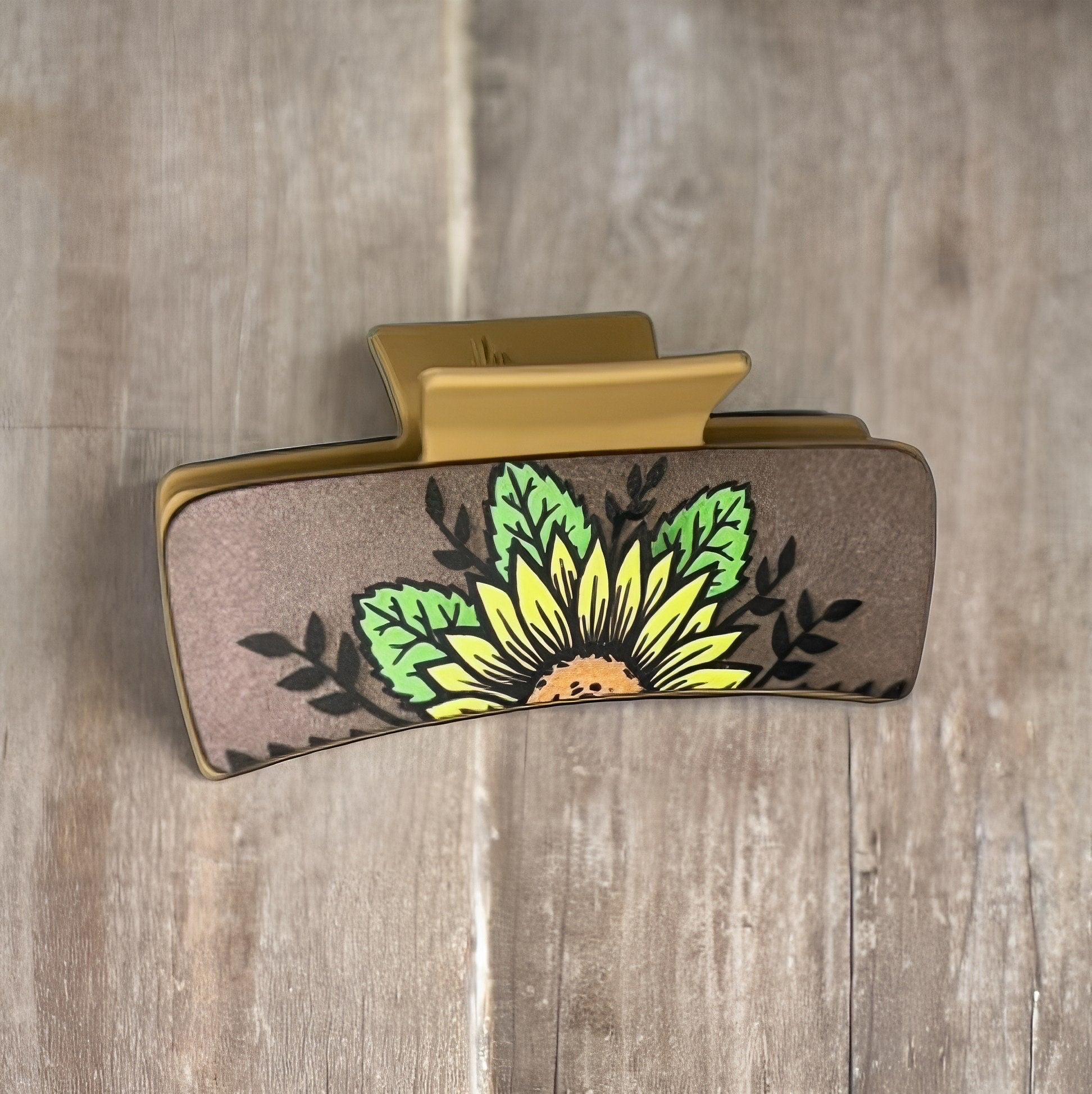 A brown colored faux leather medium shaped square claw clip with a single sunflower with green leaves and black vines in the background.