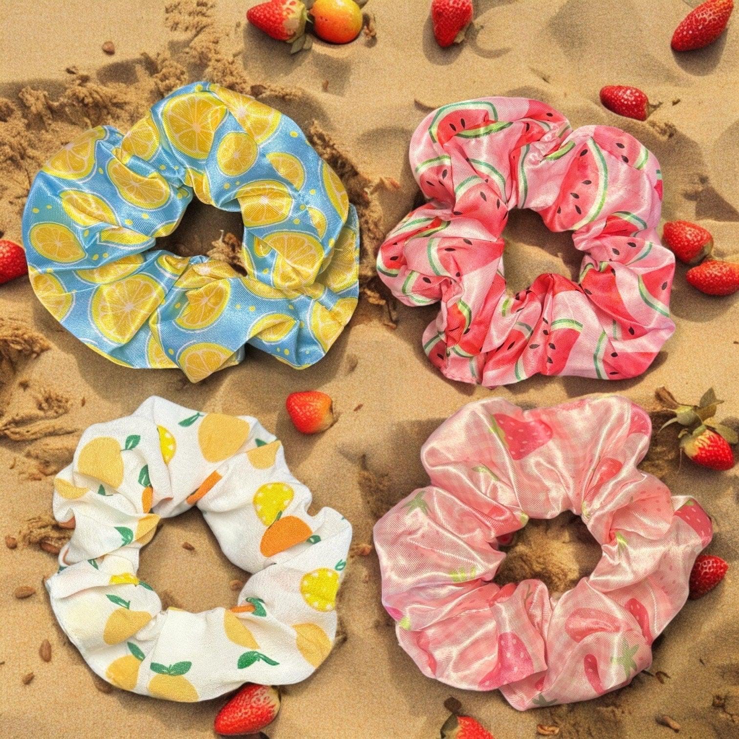 Four scrunchies with varying fruit prints. One is blue with a lemon print, one is pink with a watermelon print, one is white with an orange print, and one is pink with a strawberry print.