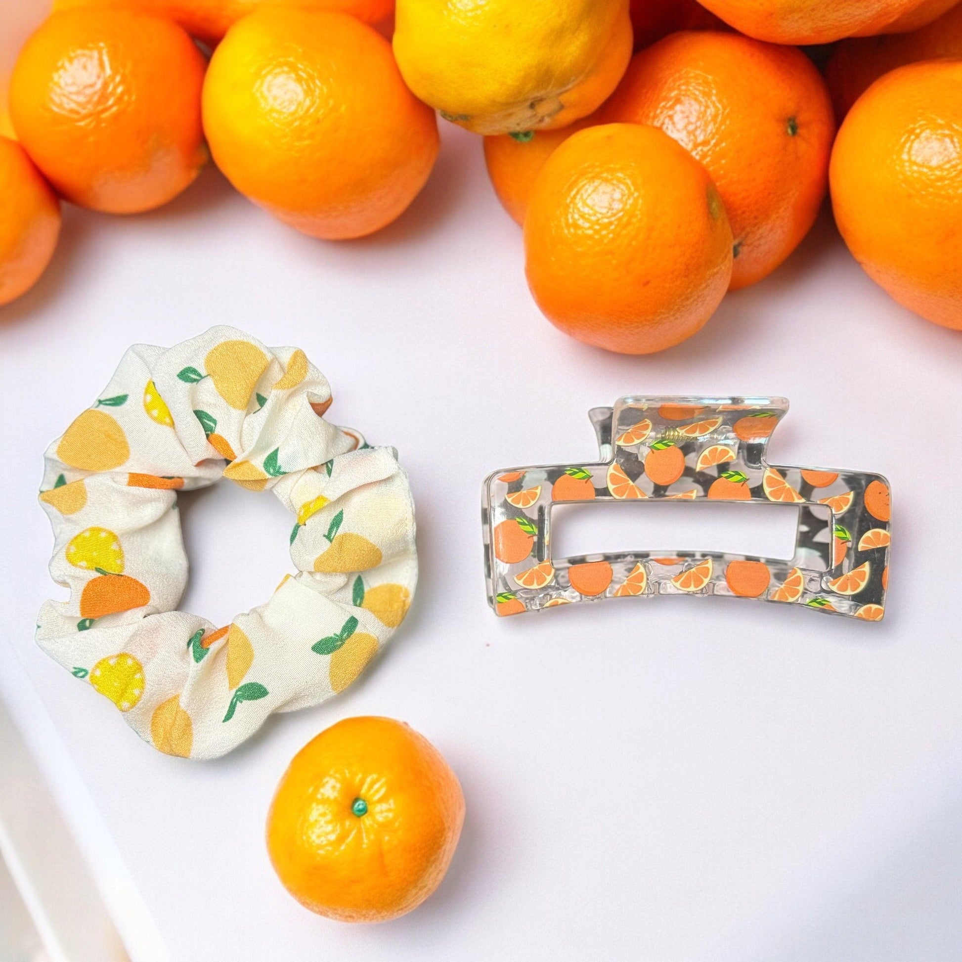 One white scrunchie with oranges on it and one clear medium square shaped claw clip with oranges printed on it on a white table with oranges in the background as scenery.