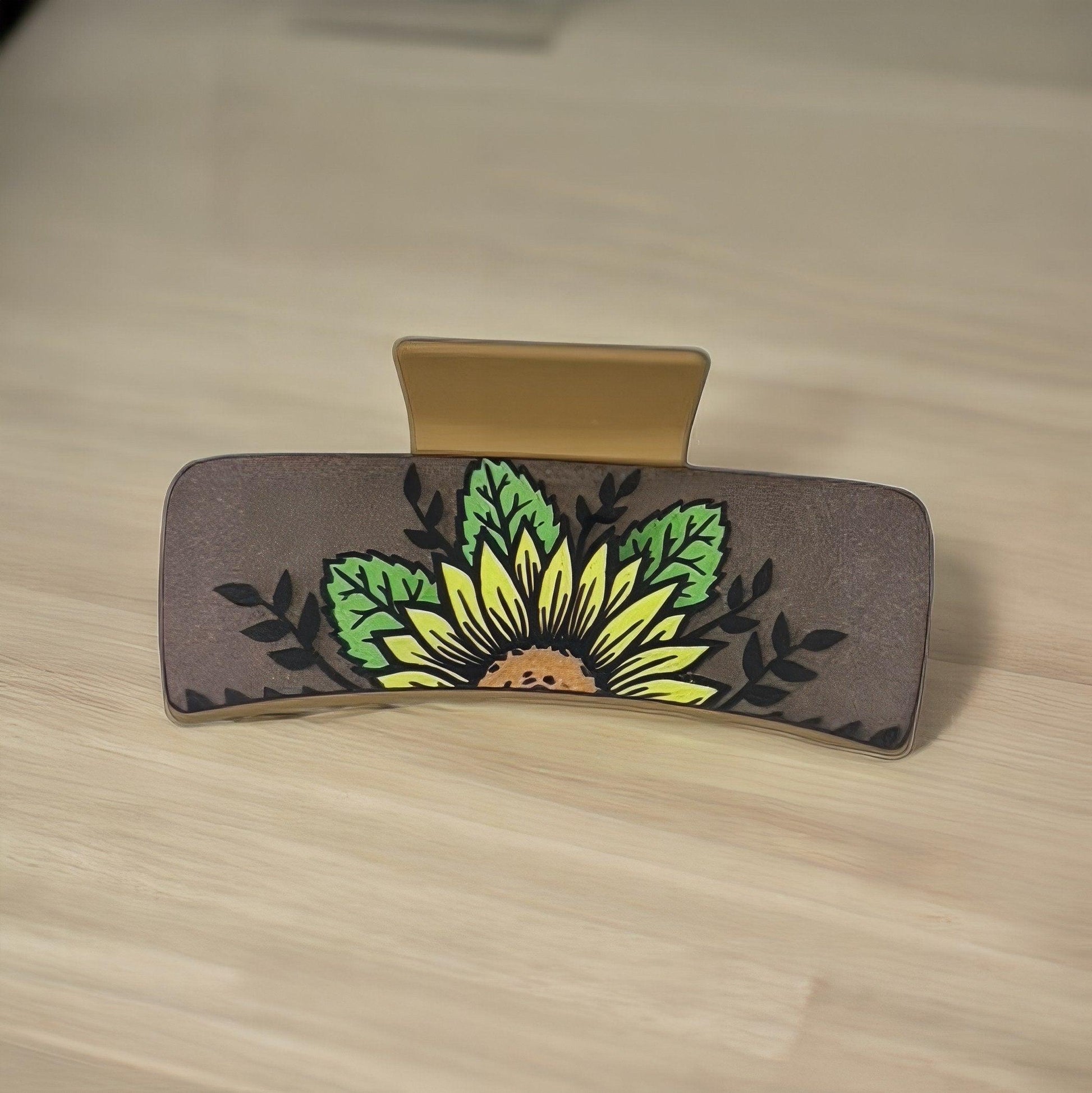 A brown colored faux leather medium shaped square claw clip with a single sunflower with green leaves and black vines in the background.