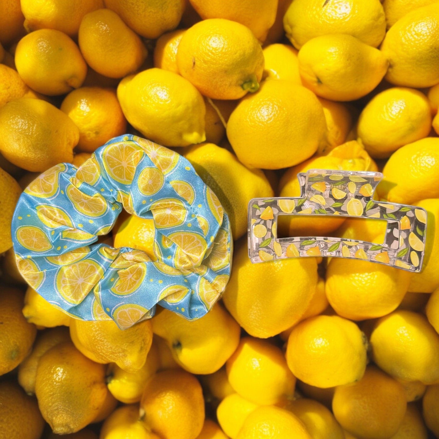 One blue scrunchie with yellow lemons printed on it with one clear medium square shaped claw clip with lemons printed on it on top of a pile of lemons as scenery.