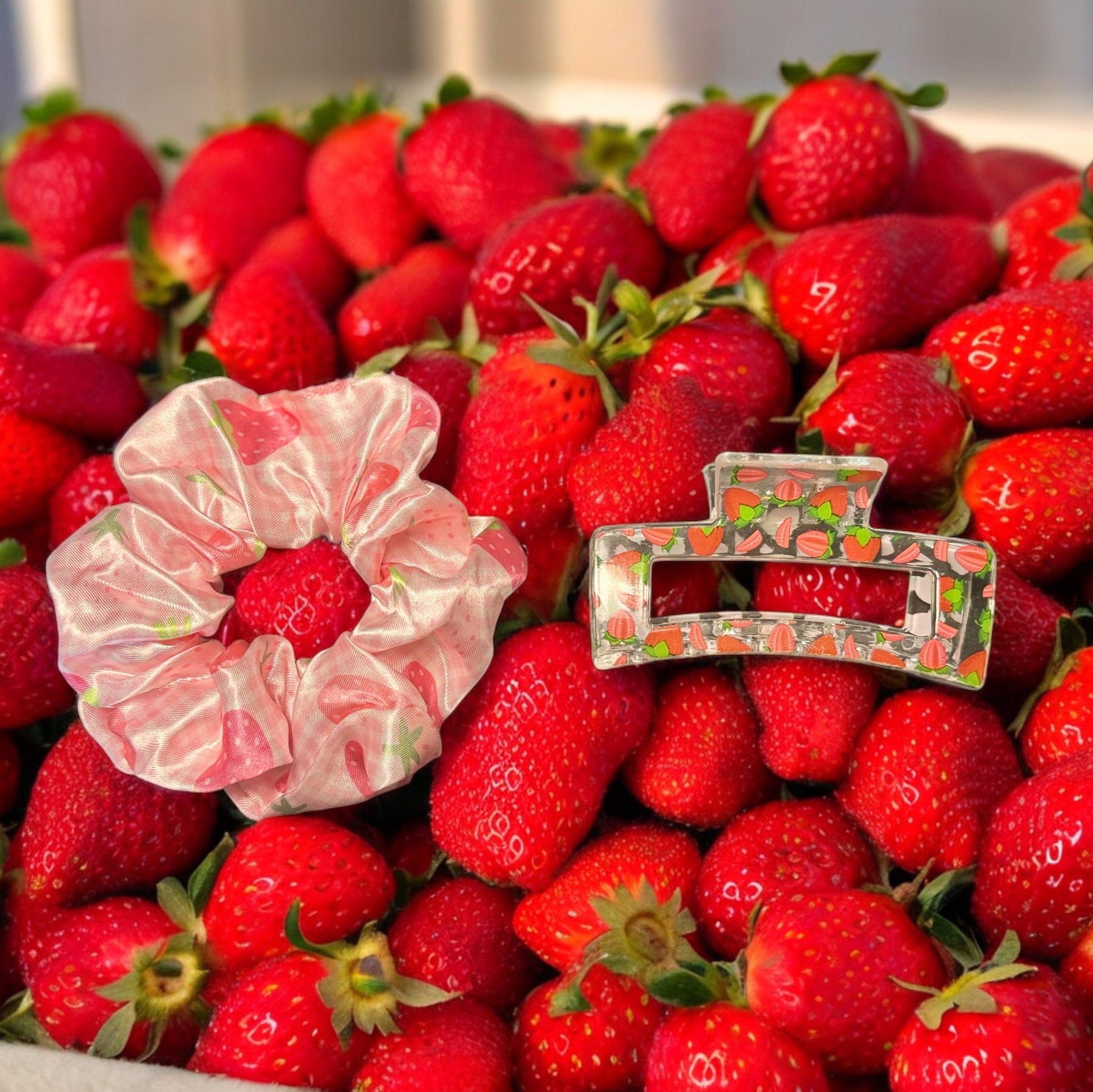 One pink scrunchie with strawberries printed on it with a clear medium square shaped claw clip with strawberries printed on it on top of a pile of strawberries as scenery.