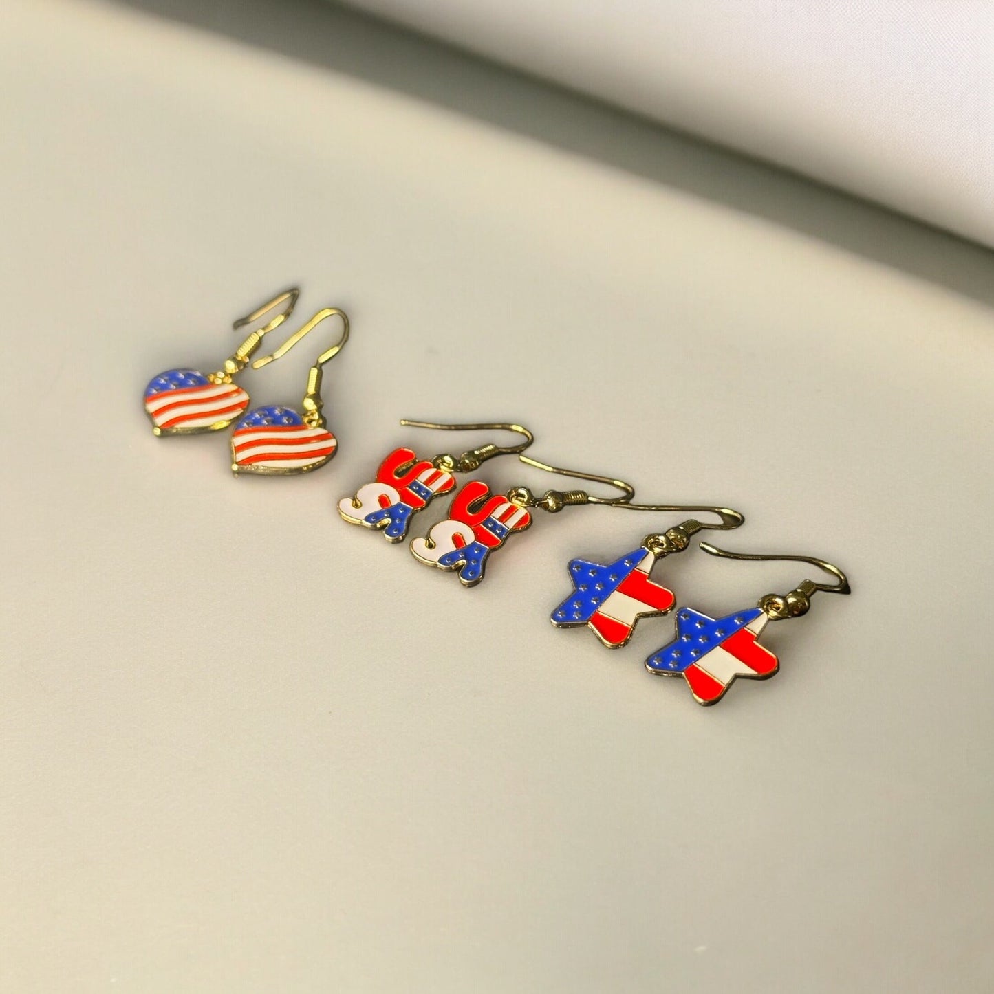 3 sets of 4th of July themed dangle earrings laying sideways. All are printed like the U.S Flag. One is heart shaped, one is shaped in bubble letters spelling USA, and one is star shaped.