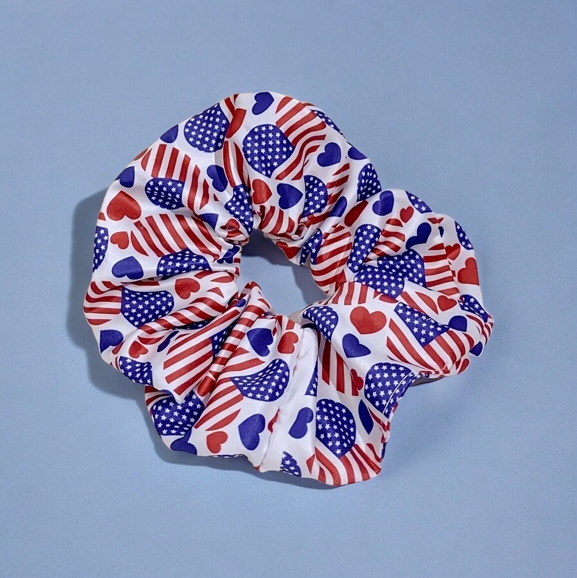 One 4th of July themed scrunchie. It is white with various red and blue colored hearts. Some of the hearts are larger with the full American flag print on them, some are smaller in colors blue and red.