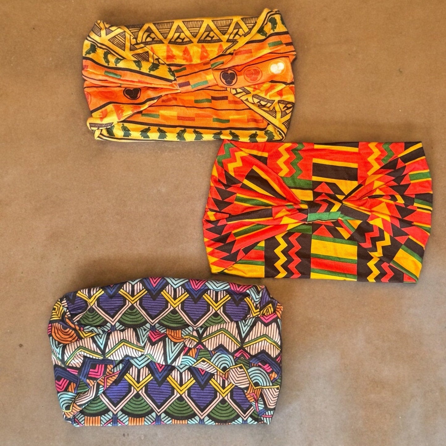 Three stretchy polyester African tribal print headbands in various colors laying on a neutral colored brown-tone background. The photo is zoomed in.