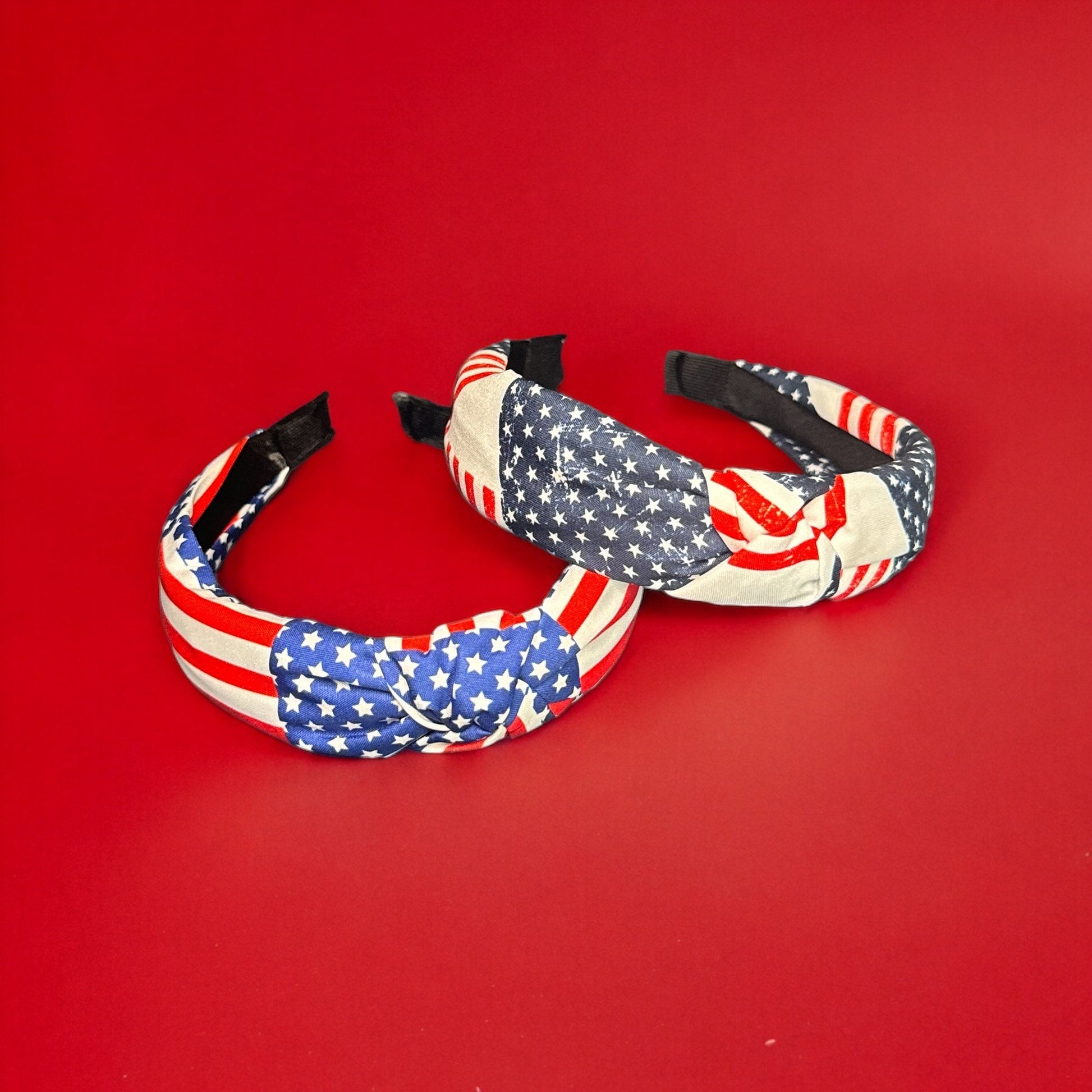 Two wide band fabric headbands with American flag prints on them laying on top of each other on a red surface. The photo is taken from an aerial view.