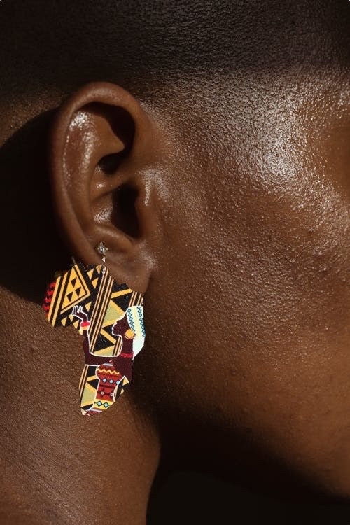 A side profile of a black woman wearing a dangle earring shaped like the continent of Africa with black and yellow tribal print and a beautiful African woman with a tribal print outfit, earrings and a head covering on the front.